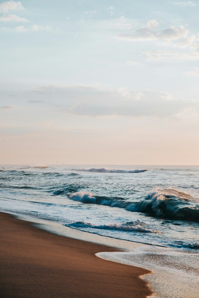 Gentle ocean waves lap against a sandy shore under a pastel sky at dawn