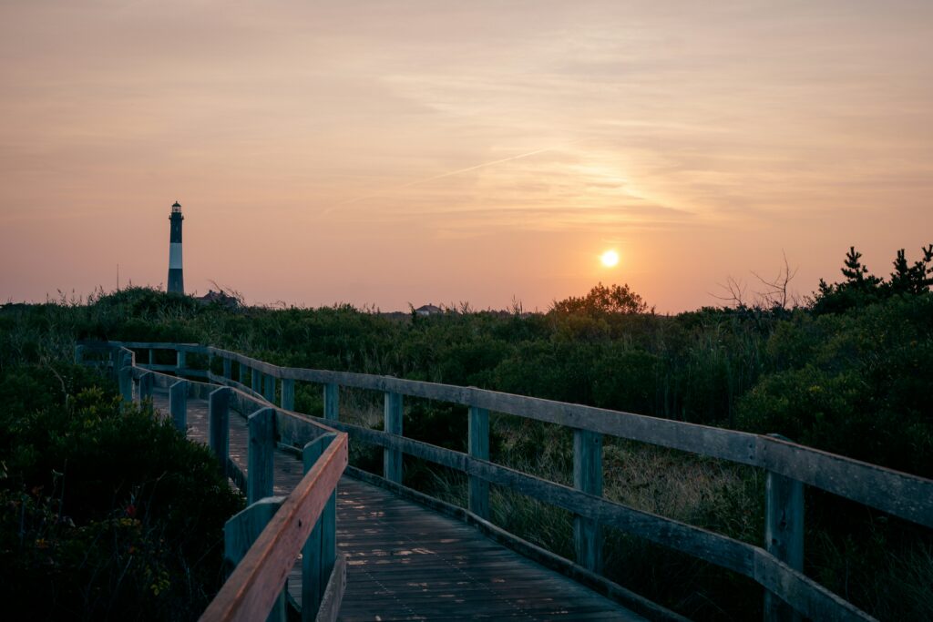 A winding wooden boardwalk with railings leads through dense coastal scrub at sunset towards a tall, striped lighthouse under a hazy orange sky.