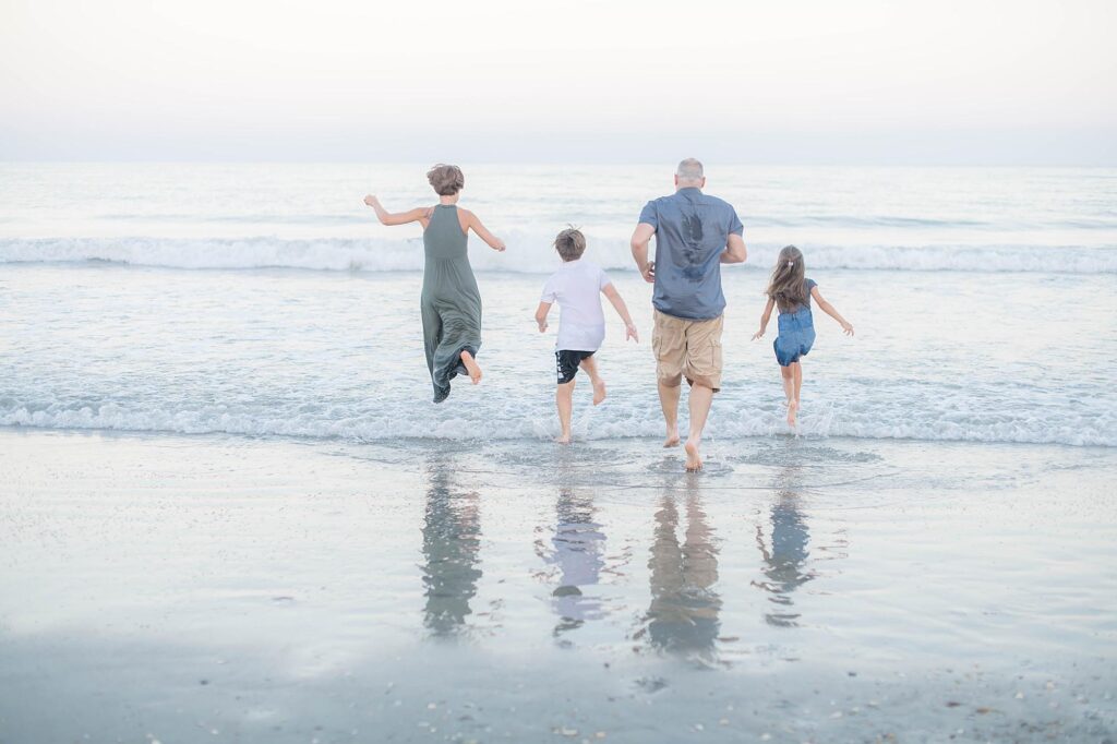 A family of four running away from the camera into the ocean waves at the beach, seen from behind
